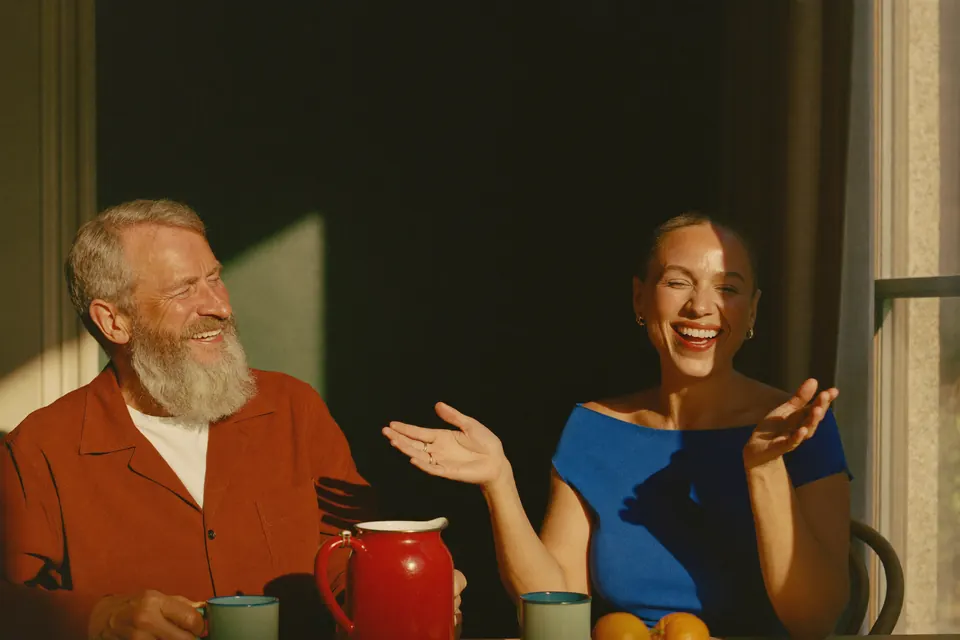 A couple in their fifties at a kitchen table in low golden sun, she is telling a story with her hands and he is laughing.