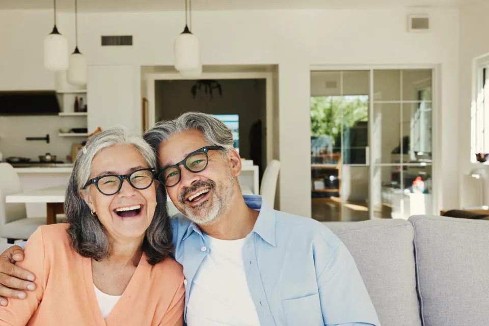 A couple in their sixties, both in glasses, sitting close on a grey sofa in a bright open plan room and laughing straight at the camera.