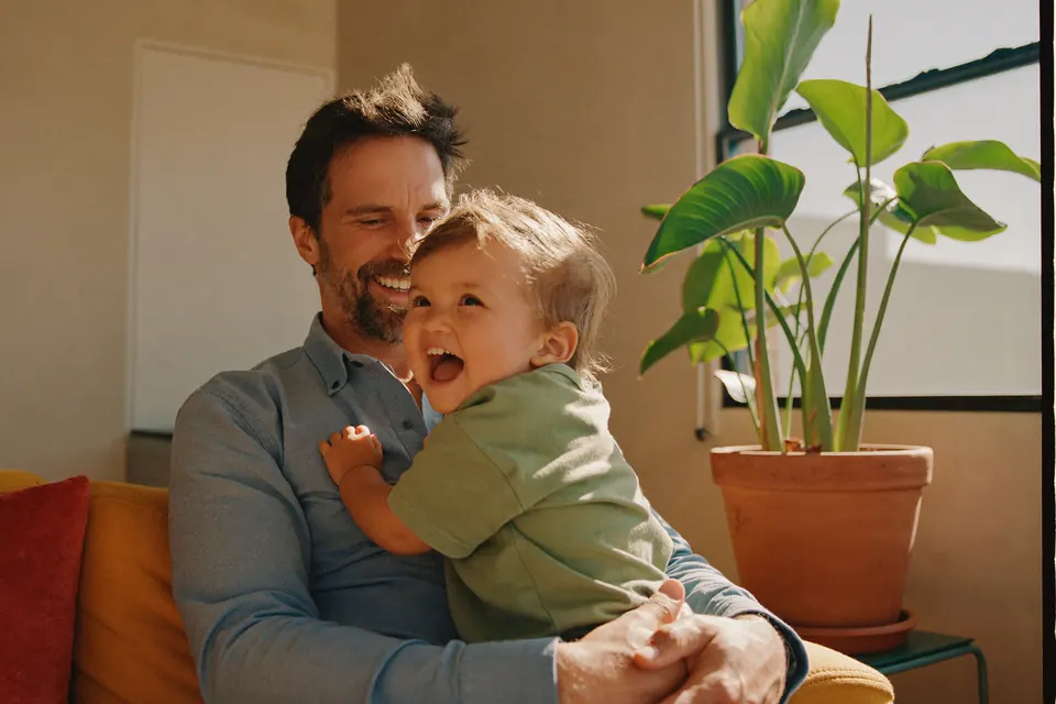 A man in his forties on a yellow sofa with a toddler on his lap, both laughing, the child reaching up to his face.
