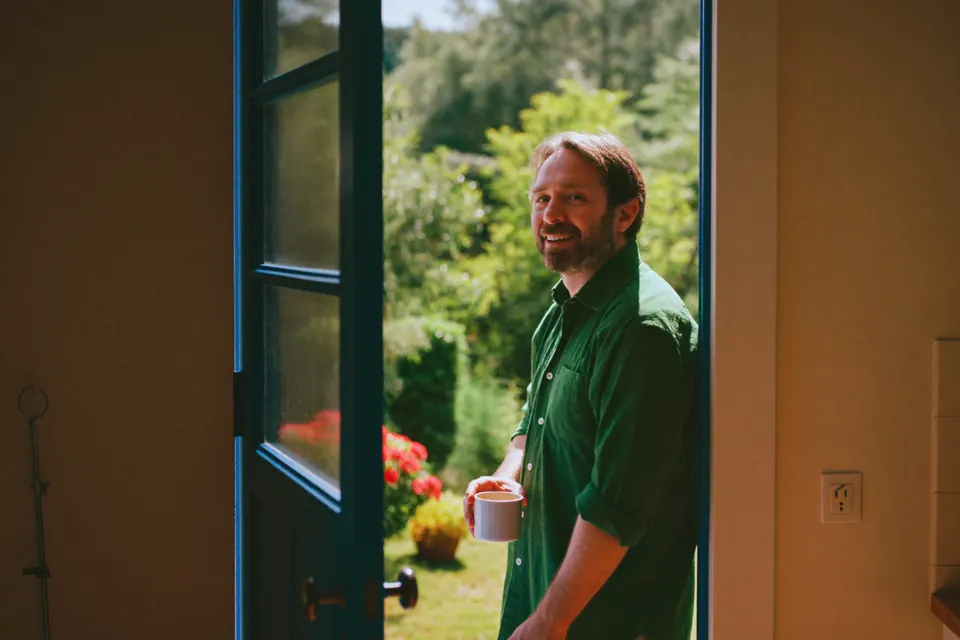 A man in the open back doorway of his kitchen with a mug, turning back into the room with a smile, a sunlit garden behind him.