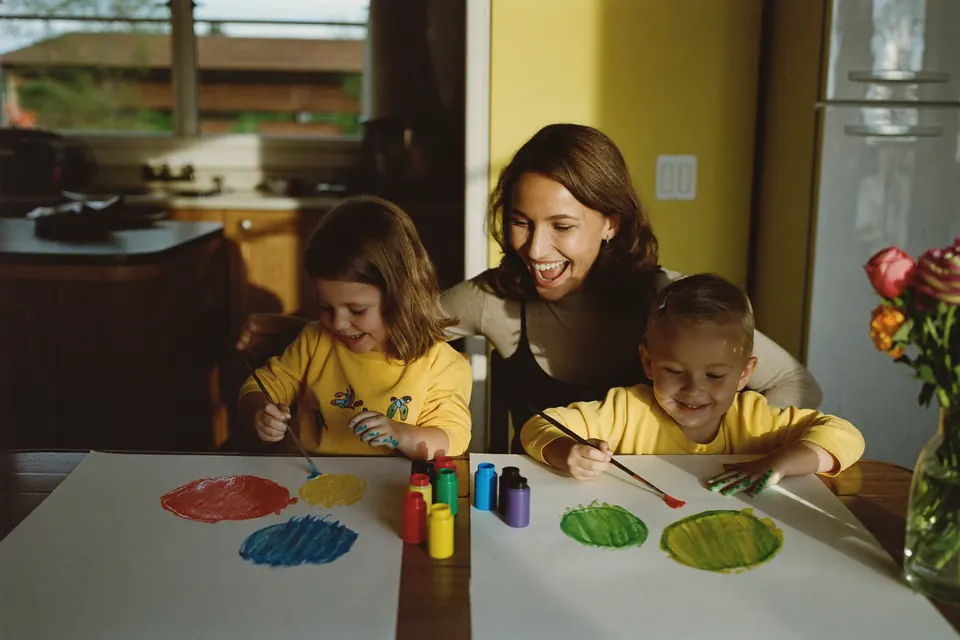 A woman laughing with her two young children at a kitchen table as they paint bright circles on big sheets of paper.