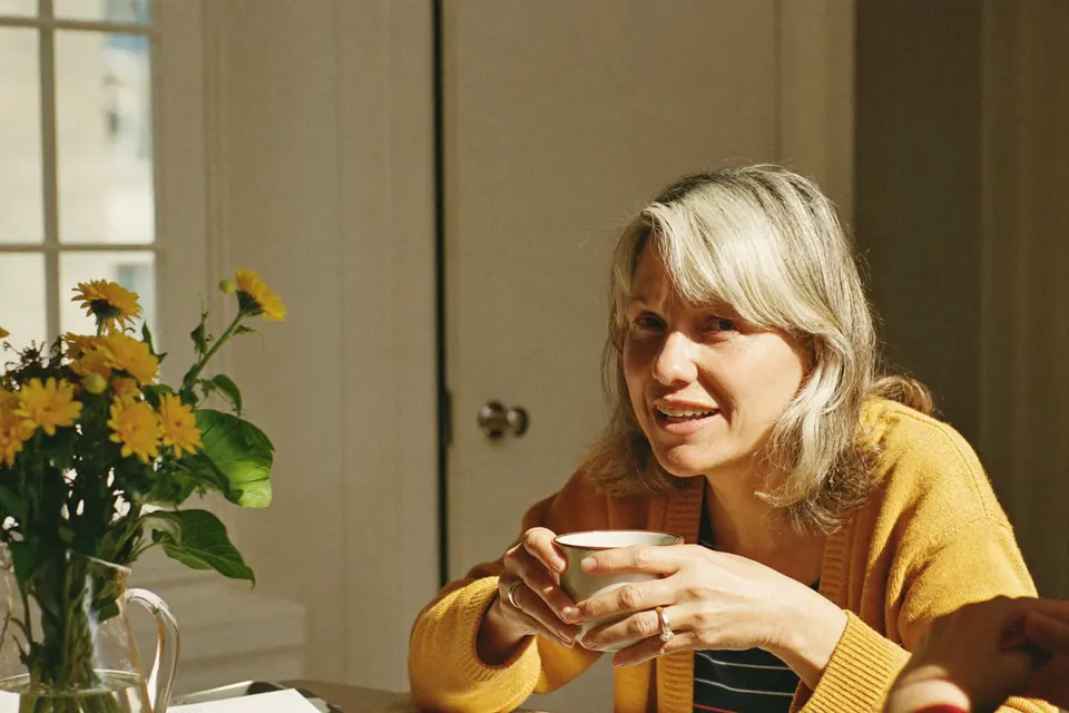 A woman in her fifties at her kitchen table in the morning sun, a mug in both hands, mid conversation with a friend across the table and looking towards the camera.