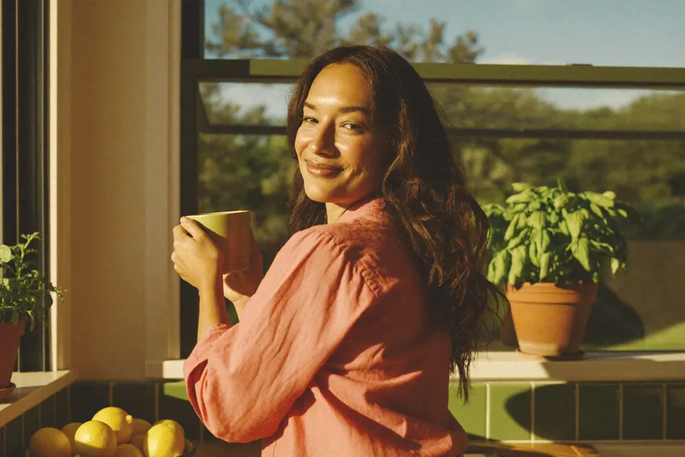 A woman in her thirties at her sunlit kitchen window with a mug, looking back over her shoulder with a smile.