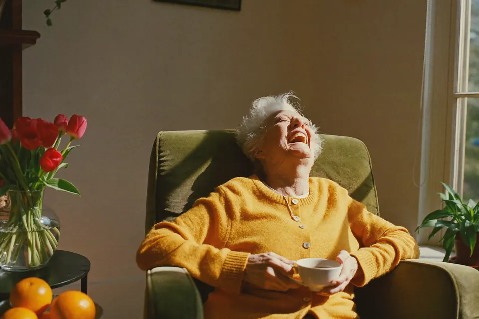 A woman in her seventies in a green armchair by a sunny window, head back, laughing, a cup of tea in her hands.