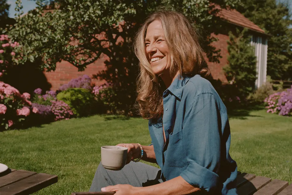 A woman in her sixties on a bench in her sunny back garden, laughing, a mug in her hand, pink flowers behind her.